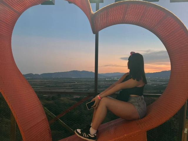 Genesiss with brown hair, pink bow, black crop top, and high-waisted shorts, sits on heart-shaped orange mesh, holding phone, wearing black-and-white sneakers, sunset in background.