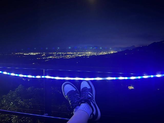 Genesiss's legs with black and white sneakers, blue laces, and white socks, resting on a glass railing, under a dark night sky with city lights below.