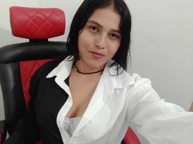 ValentinaMoreau with black hair, white shirt, black vest, showing cleavage in lace bra, sitting in red and black chair, wearing black bead necklace, smiling slightly.