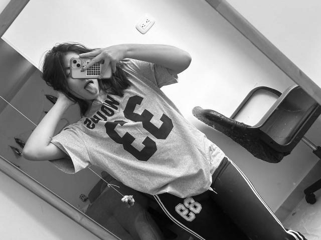 SpencerLe with a messy face, tongue out, wearing a loose gray "Broncos 53" shirt and dark pants, taking a mirror selfie in a room. Black-and-white photo.