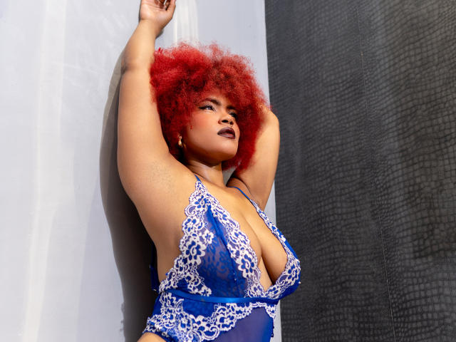 Alice Jelanni mit roten, lockigen Haaren, Armen erhoben, trägt ein blaues Spitzendessous-Kleid und präsentiert damit ihre großen, auffälligen Brüste. Sie hat dunkelroten Lippenstift und einen selbstbewussten, sexy Ausdruck..