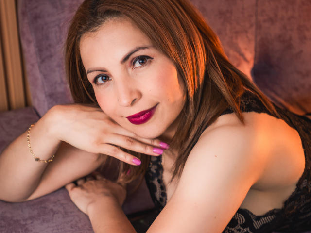 VivienneNoir, con cabello castaño, lápiz labial púrpura y uñas rosas, acostada en un sofá púrpura, vestida con encaje negro, sonriendo, con una mano en la mejilla, una pulsera dorada, luz naranja suave..