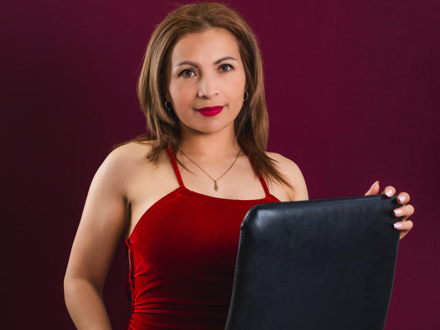VivienneNoir with brown hair, red dress, holding black leather chair, wearing red lipstick and necklace, smiling slightly, in a studio shot.