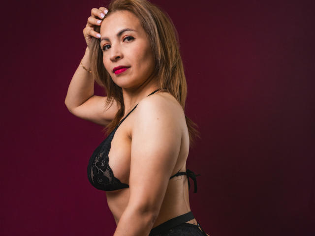 VivienneNoir, con cabello castaño, lápiz labial rojo, un sujetador de encaje negro y tanga a juego, con la mano en el cabello, vista de perfil, sonriendo con confianza, busto medio, delgada, piel clara, fondo rojo oscuro..