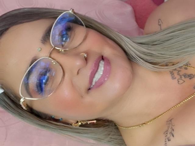 AlessadraJoness heeft blauwe oogschaduw, een bril, een neusring, lipgloss, tatoeages, blond haar, een gouden ketting en een vrolijke glimlach. Borsten en billen zijn niet zichtbaar..