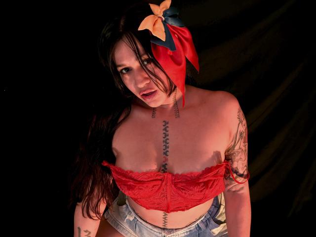 AbbyMontgomery with long black hair, red lace top, small tits, tattoos, and a red hat with a yellow bow, looking up with an open mouth, wearing denim shorts.