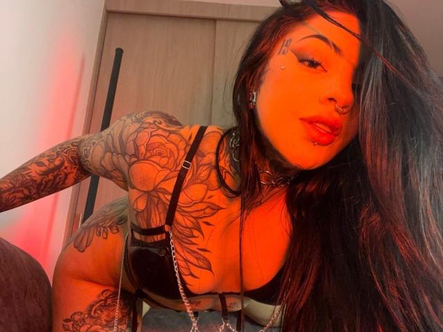 Milamonty with orange glow, black lingerie, and tattoos, leaning forward, showing tits and rose tattoo on arm, long black hair, nose ring, and red lipstick.