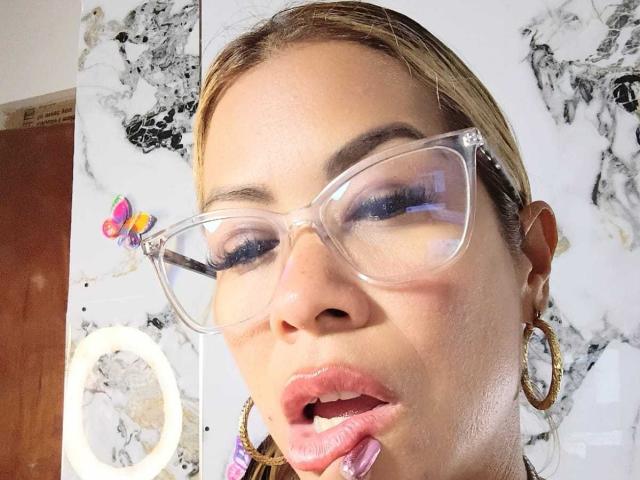 AlixLow, avec des lunettes transparentes, des lèvres roses brillantes et des boucles d'oreilles cercles, tenant une brosse à dents rose dans sa bouche, portant une pince à cheveux colorée, regardant légèrement sur le côté..