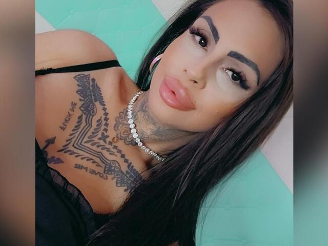 DenyChantal has dark hair, thick eyebrows, and full pink lips. She's wearing a black top, showing off her tattoos and necklace. Her tits are partially visible.