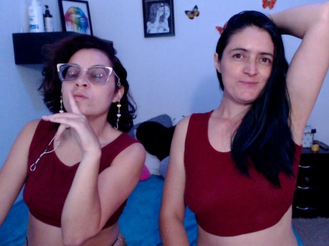 LunaVenus com óculos, lábios curvados e brincos, e LunaVenus com cabelo preto comprido, braço levantado, ambas usando tops vermelhos, mostrando seus seios em um quarto..