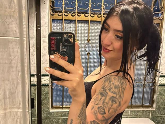 TifanyDiaz, avec ses cheveux foncés attachés en queue de cheval, portant un soutien-gorge en dentelle noire, tenant un téléphone, et présentant un tatouage détaillé du visage d'une femme sur son bras droit..