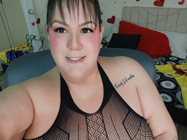 GiiannaMorrys with pale skin, dark hair, and pink eyeshadow, wearing a black fishnet top, showing a tattoo on her right shoulder, smiling at the camera.