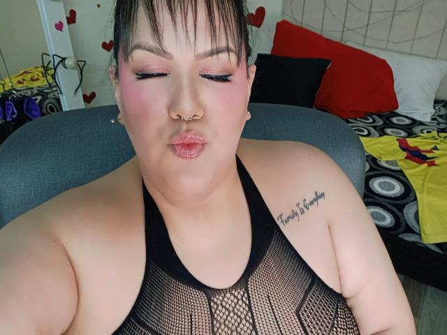 GiiannaMorrys with pink eyeshadow, nose ring, and black mesh top, pouting lips, tattoo on right shoulder, short dark hair, sitting on gray chair, showing her bare tits.