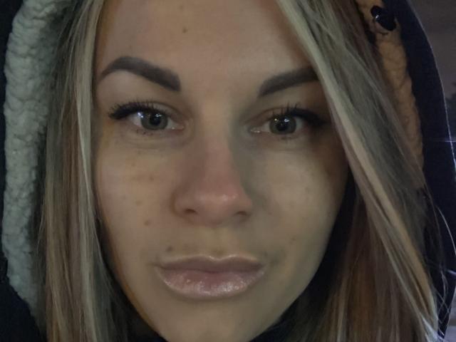 BelleLisaG ha i capelli biondi, le sopracciglia scure e indossa una giacca con cappuccio e un colletto peloso. Il suo viso è leggermente lucido, con pori visibili e piccole macchie di acne..