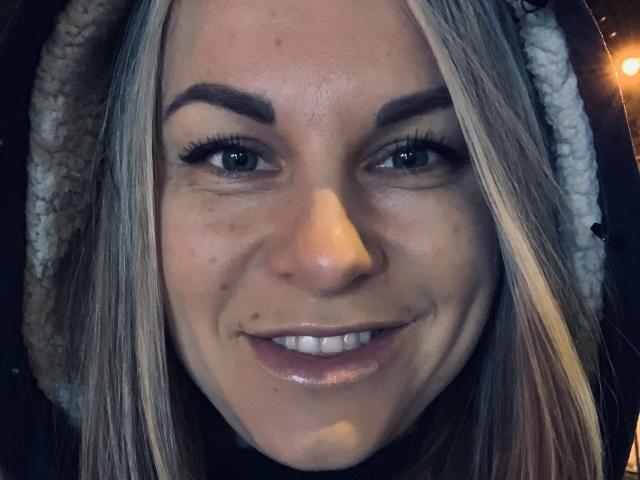 BelleLisaG ha i capelli biondi, gli occhi azzurri e un leggero sorriso con labbra rosa lucide. Indossa un cappotto nero con cappuccio e fodera in pile bianco..