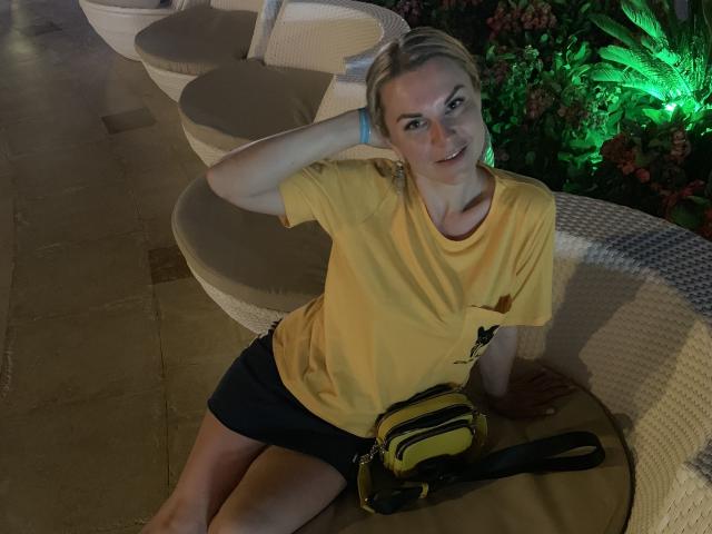 BelleLisaG avec des cheveux blonds, un t-shirt jaune, un short noir, un sac jaune et noir, et une laisse, assise sur des chaises en osier blanc, une main dans les cheveux, des plantes éclairées en vert en arrière-plan, souriante..