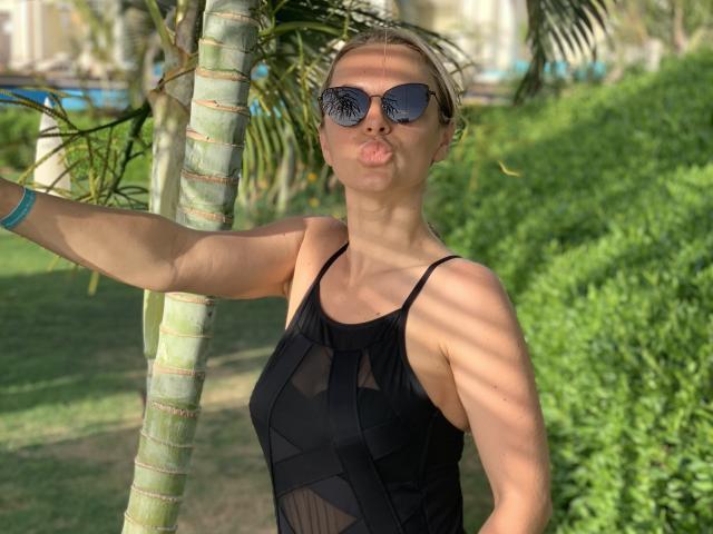 BelleLisaG avec des lèvres pulpeuses, portant un haut noir transparent, des lunettes de soleil, les cheveux blonds attachés, un bras autour d'un palmier, un bracelet bleu, la lumière du soleil projetant des ombres..