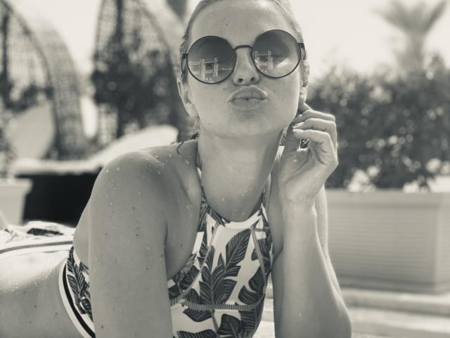 BelleLisaG avec des lunettes de soleil rondes, des lèvres pulpeuses et un haut de bikini fleuri, des gouttes d'eau sur sa peau, une main près de son oreille, penchée en avant, en noir et blanc, dans un décor tropical..
