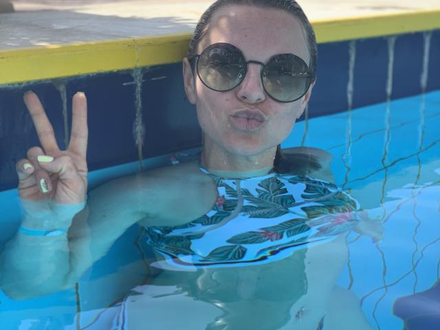 BelleLisaG avec des cernes visibles derrière ses lunettes de soleil, des lèvres boudeuses, un haut floral mouillé et des ongles néon, faisant un signe de la paix dans une piscine, montrant ses seins et ses fesses sous l'eau..