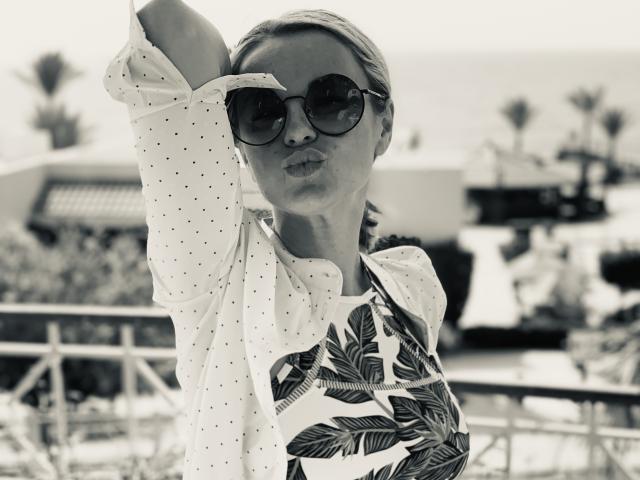 BelleLisaG avec des lunettes de soleil, une chemise bouffante et un haut à motifs de feuilles, faisant un baiser, levant le bras, cheveux blonds, arrière-plan flou, photo en noir et blanc, sourire discret, pose confiante..