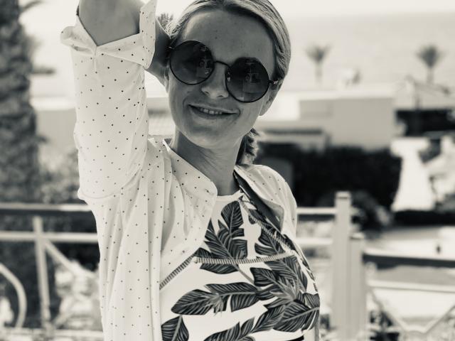 BelleLisaG avec des lunettes de soleil, souriante, levant le bras, portant une chemise blanche à pois avec un motif de feuilles, cheveux blonds en tresse, tenant un sac à main, debout à l'extérieur, photo en noir et blanc..