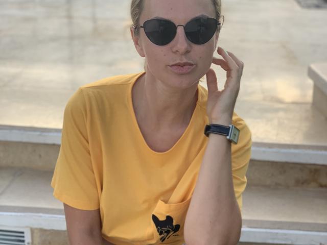BelleLisaG, avec des cheveux blonds, portant des lunettes de soleil foncées, un t-shirt jaune et une montre noire, touchant son oreille avec sa main droite, avec une expression légèrement boudeuse..
