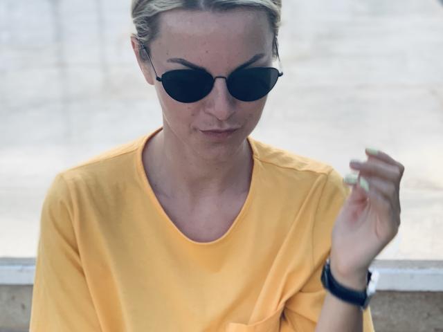 BelleLisaG avec des cheveux blonds, des lunettes de soleil noires, une chemise jaune et un bracelet noir, tenant une cigarette, avec un léger sourire, regardant vers le bas, la main droite levée..