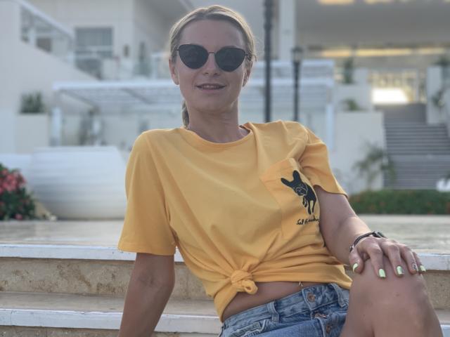 BelleLisaG, t-shirt jaune noué à la taille, short en jean, lunettes de soleil noires, cheveux blonds attachés en queue de cheval, ongles verts, assise sur les marches, souriante, tatouage d'un chien noir sur le t-shirt..