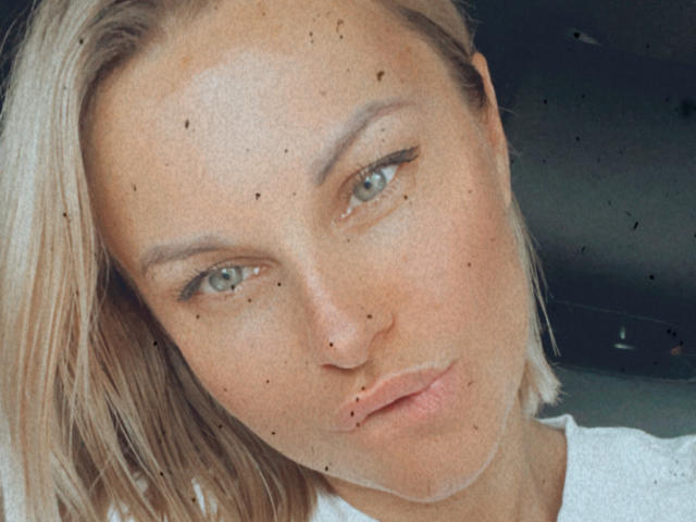 BelleLisaG a les cheveux blonds, les yeux bleus et un visage légèrement boudeur avec de petites taches de rousseur. Elle porte une chemise blanche, et son maquillage comprend un trait de crayon noir fin..