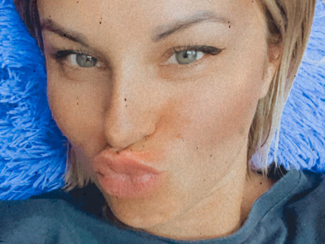 BelleLisaG, avec ses lèvres pulpeuses, sa peau pâle, ses yeux marron clair et ses cheveux blonds, portant une chemise bleue, faisant une moue de baiser, a de petites taches de rousseur sur son visage..