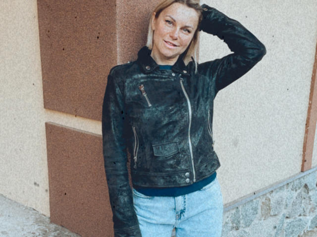 BelleLisaG, portant une veste en cuir noir usée et un jean bleu, se tient contre un mur, une main dans les cheveux, souriant légèrement, montrant de petites seins et une expression mignonne et détendue..