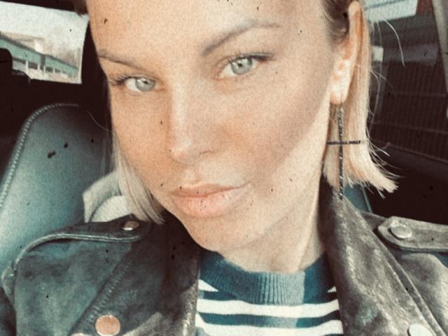 Le visage boudeur de BelleLisaG, ses yeux verts et ses courts cheveux blonds. Elle porte une chemise rayée et une veste en cuir vert avec des boutons. Ses boucles d'oreilles sont petites et métalliques..