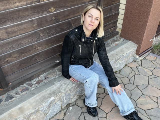 BelleLisaG, blonde, assise sur des marches de pierre, portant une veste en cuir noir, un jean bleu et des bottes noires, regardant l'appareil photo, décontractée, détendue, vue de face, en extérieur, en journée, confiante..