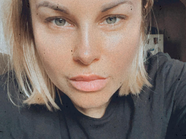Le visage de BelleLisaG, avec ses cheveux blonds mi-longs, ses yeux verts et ses lèvres roses légèrement entrouvertes. Elle porte une chemise noire, avec quelques petites taches sombres sur sa peau..