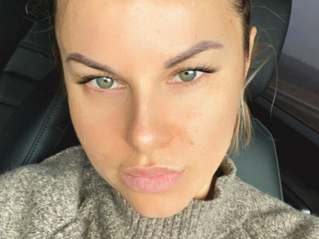 Le visage de BelleLisaG, avec ses yeux verts, ses sourcils épais et ses lèvres roses légèrement gonflées, porte un pull gris, assise sur le siège en cuir noir d'une voiture. Sa peau présente de petites imperfections..