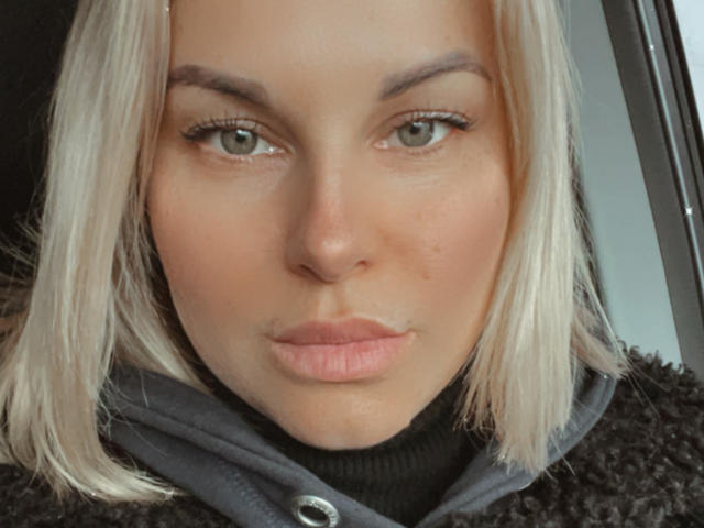 Le visage de BelleLisaG, peau pâle, yeux verts, coupe de cheveux blonde, légère moue, portant un sweat à capuche noir texturé, maquillage discret, petit nez, petites lèvres, joues légèrement rosées, regardant directement l'appareil photo..