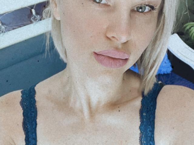 BelleLisaG, avec sa peau pâle, ses cheveux blonds et ses lèvres roses, porte un soutien-gorge en dentelle bleu, mettant en valeur son cou fin et sa clavicule. Ses seins sont petits et ses yeux sont bleus..