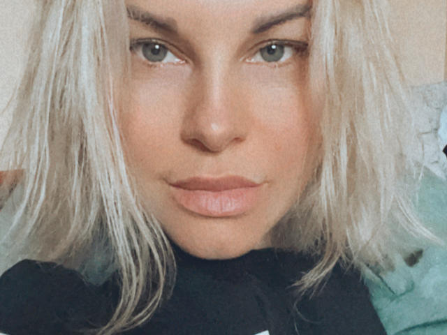 Le visage de BelleLisaG, mettant en valeur ses lèvres pulpeuses et ses yeux verts, avec ses cheveux blonds platine et un léger sourire, sur un selfie avec un effet de flou doux..