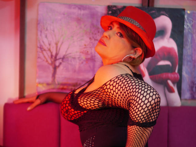BellaMystic com chapéu vermelho, blusa de meia-calça e brincos grandes, mostrando os seios, com uma luz rosada e um olhar sexy e confiante, em pé em frente a um sofá rosa..