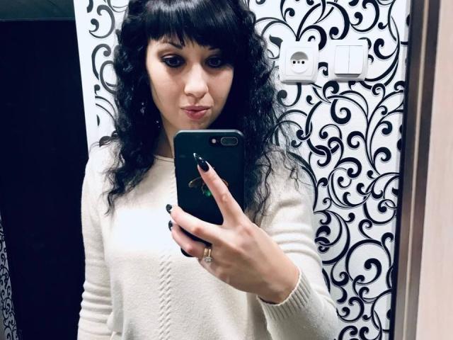 LangoriaJoy mit lockigem, schwarzem Haar, die einen weißen Pullover trägt und ein Selfie vor einem Spiegel mit ihrem Handy macht, mit schwarzem Nagellack und einem Ring am Finger, sieht süß und selbstbewusst aus..