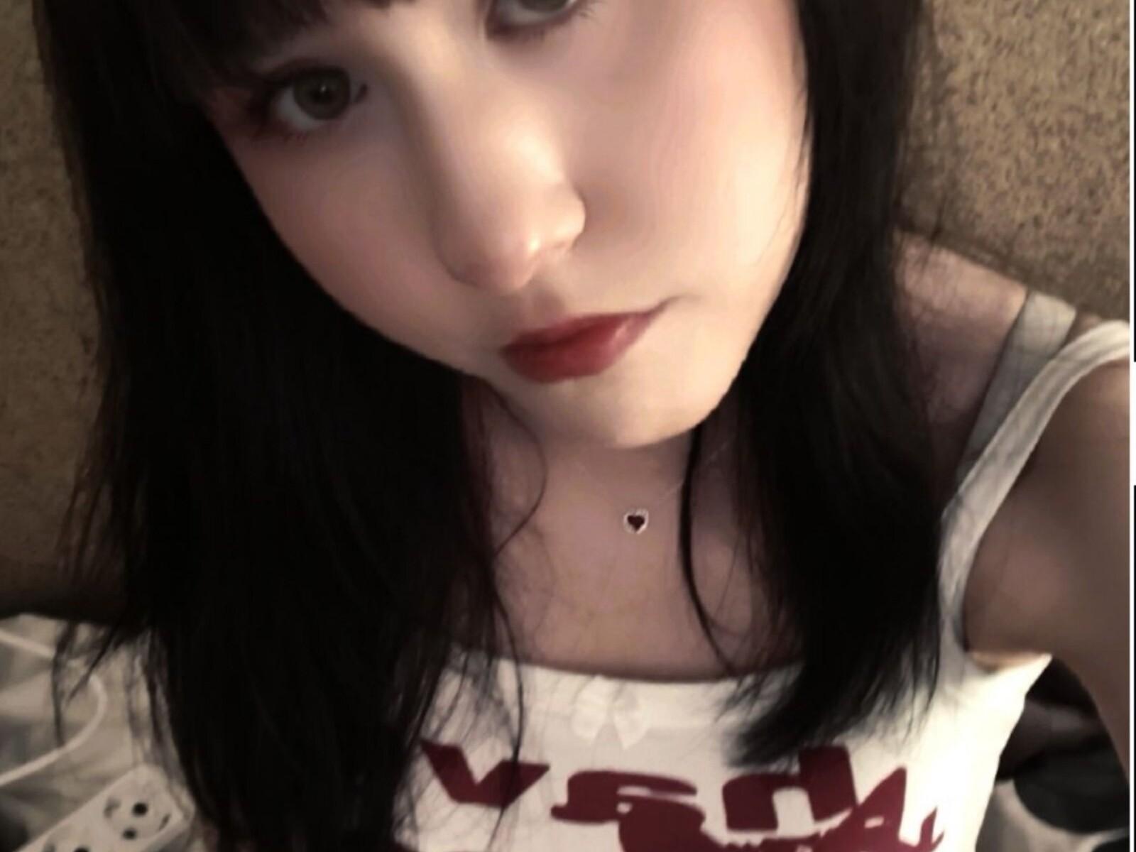JamaSlomba's pale face with dark, straight hair, red lipstick, green eyes, and white tank top, showing a small black heart necklace. Boobs not visible.