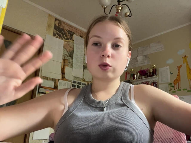 KrisEmpey, avec des cheveux bruns clairs, portant un débardeur gris côtelé, des écouteurs blancs, et une expression neutre, légèrement boudeuse, avec une main levée au premier plan..