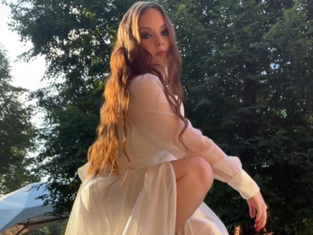 KrisEmpey, avec de longs cheveux bruns ondulés, une robe blanche transparente, accroupie, regardant par-dessus son épaule, montrant ses fesses, dans un décor extérieur ensoleillé, entourée d'arbres verts..
