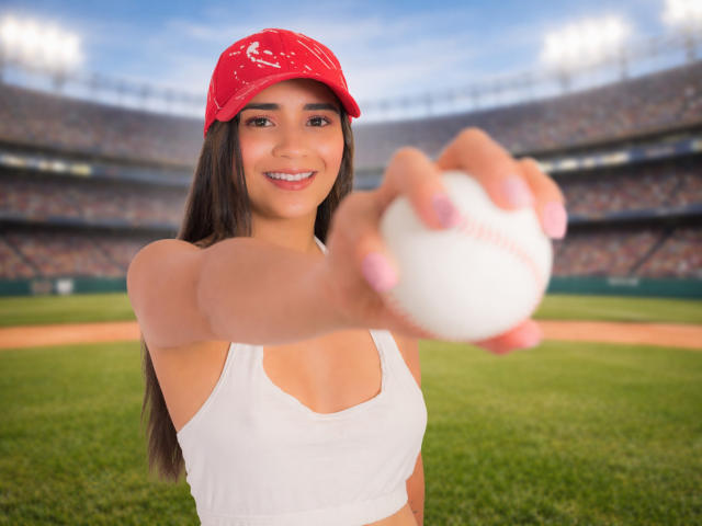 LunaBrittonn, avec de longs cheveux bruns, une casquette rouge, un soutien-gorge de sport blanc et des ongles roses, lance une balle de baseball blanche, en souriant, dans un stade lumineux. Ses seins sont légèrement visibles..