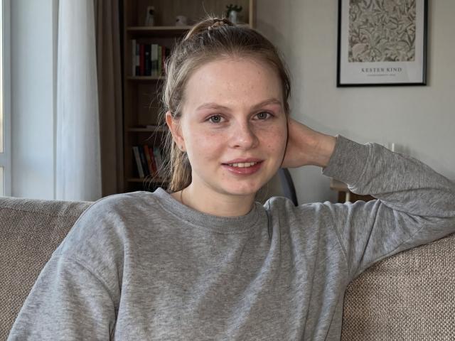 RendaKu mit Sommersprossen, graues Langarmshirt, braunes Haar im Pferdeschwanz, Hand hinter dem Kopf, leichtes Lächeln, auf einem beigen Sofa, natürliches Licht, dezentes Make-up, lässig, entspannt, attraktiv..
