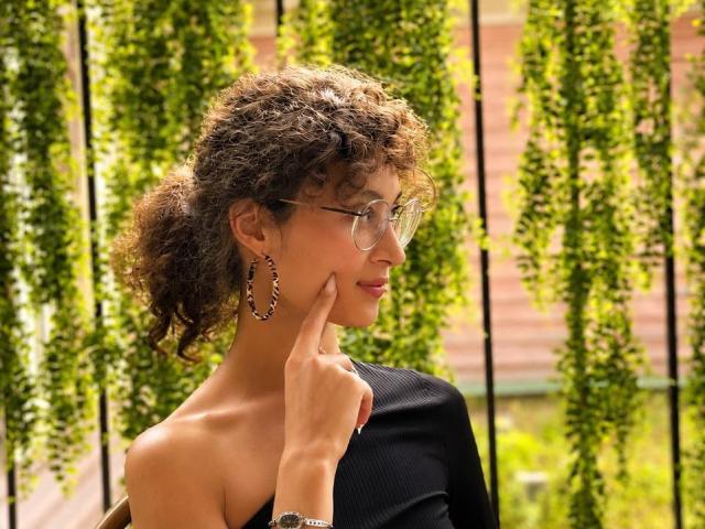 AlyceErway with curly brown hair, glasses, and hoop earrings, touching her cheek with a finger, wearing a black top, in a sunny, green, vertical plant-filled setting.