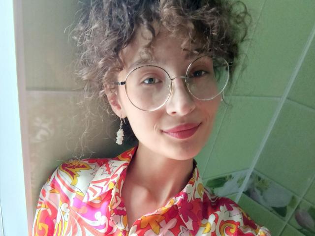 Alyce Erway, con i capelli castano ricci, indossa occhiali rotondi, una camicia colorata con fiori e un orecchino, sorride con il rossetto rosso, mostrando il suo viso carino e il suo piccolo naso..