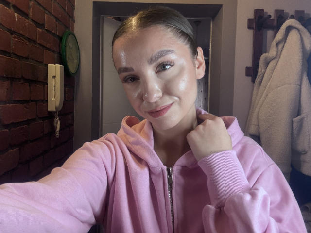 Jamalla, avec une peau éclatante, porte un sweat à capuche rose zippé, ses cheveux bruns sont attachés en arrière, et elle sourit à l'appareil photo, sa main droite est près de son cou. Un mur de briques et un téléphone sont visibles en arrière-plan..