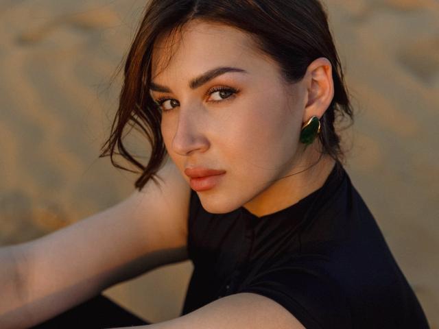 Olivia Cluff a les cheveux brun foncé, porte des boucles d'oreilles vertes et est vêtue d'une chemise noire. Elle est assise avec une expression légèrement sérieuse et séduisante, et porte un maquillage doux et naturel..