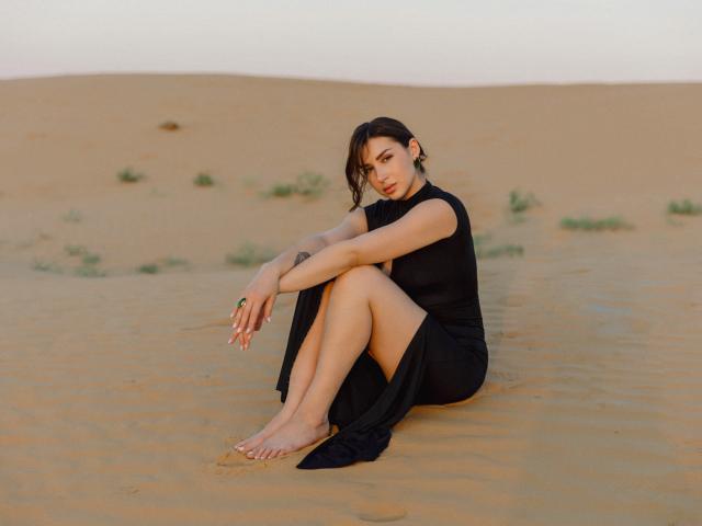 Olivia Cluff sitzt im Sand, die Beine gekreuzt, die Arme verschränkt, trägt ein schwarzes Kleid, ist barfuß, hat kurze, dunkle Haare, ein dezentes Tattoo am rechten Unterarm, weiches Abendlicht, ein ruhiger Gesichtsausdruck..